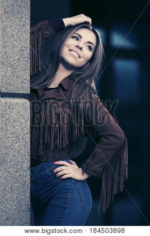 Happy young woman standing at the wall in city street. Stylish fashion model in leather fringe suede jacket and blue jeans