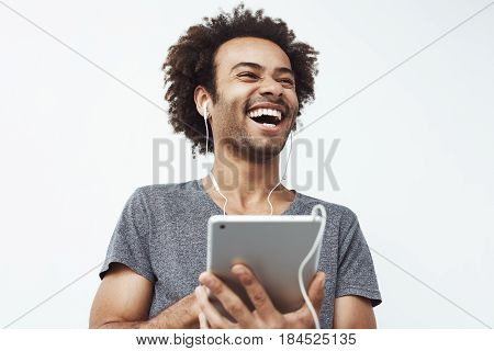 Portrait of cheerful happy african man in headphones laughing holding tablet talking or watching and enjoying a comedy show or browsing over white background.Copy space.
