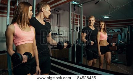 Man and woman stand near the mirror with dumbbells in the gym.