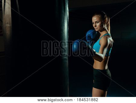 Girl boxer in blue boxing gloves near a punching bag.