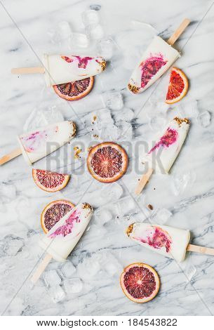 Healthy summer dessert. Blood orange, yogurt and granola popsicles on ice cubes over light marble background, top view. Clean eating, dieting, weight loss concept