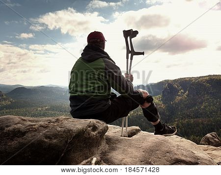 Tired Tourist With Medicine Crutch  And Broken Leg Fixed In Immobilizer Resting On Summit.