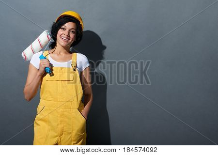 Happy smiling mixed race construction woman wearing yellow protect helmet and overall adjusting helmet holding a painting roller over shoulder standing at grey wall looking at camera.