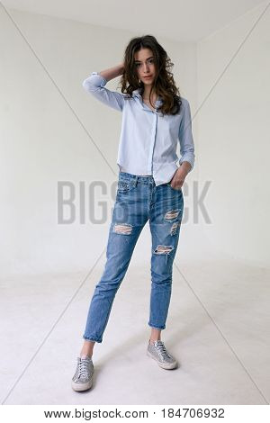 Young brunette in a blueshirt blue ragged jeans and sneakers stands in a relaxed pose on a white background