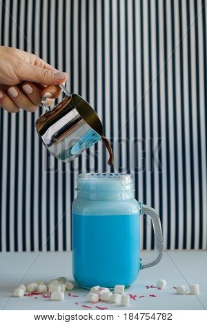 Woman is pouring coffee in cup of colored blue milk on a black-and-white background. Milk shake cocktaill frappuccino. Unicorn coffee unicorn food.