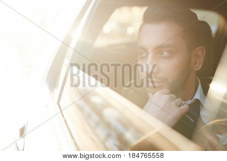 Sunlit portrait of handsome Middle-Eastern businessman riding in backseat of car  and looking out of window watching city streets