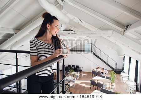 Portrait of woman standing with a cup and looking down at the people on the other floor. Happy adult woman looking away from camera