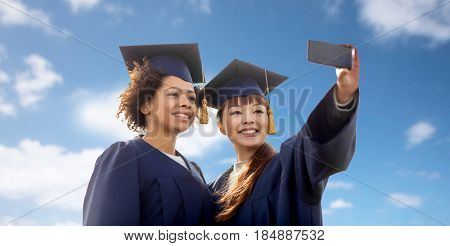 education, graduation, technology and people concept - group of happy international students in mortar boards and bachelor gowns taking selfie by smartphone over blue sky and clouds background