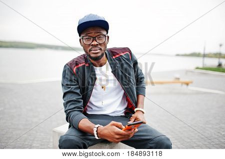 Portrait Of Stylish African American Man On Sportswear, Cap And Glasses Sitting At Stone Cube With P