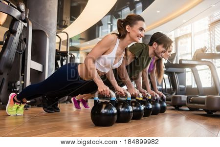 Side view of three young people smiling while doing the kettlebell plank challenge for strong core during group workout at the gym