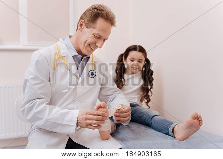 Solid fixator. Emotional handsome experienced rheumatologist treating his little patient and applying special medical material while she sitting on a bed in his office