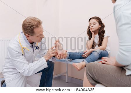 Special spots. Competent skillful professional rheumatologist examining little patients feet and explaining the diagnose to her mom while the girl sitting on a bed