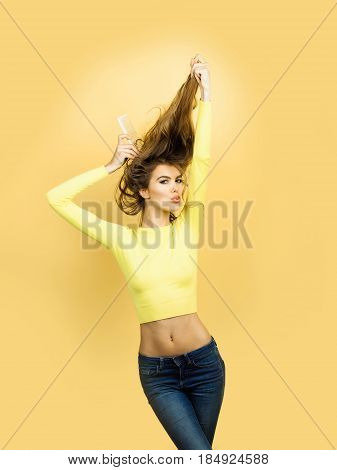 girl or pretty woman fashion model in stylish yellow top with sexy belly combing wavy blond long hair with comb on orange background. Hairdressing and beauty salon