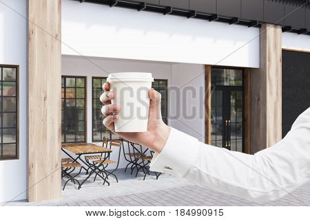 Close up of a businessman s hand holding a coffee to go near a cafe with gray walls and two wooden tables with chairs standing near a door. 3d rendering