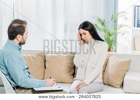 Unhappy Young Woman With Depression Sitting On Couch And Psychiatrist Writing On Clipboard