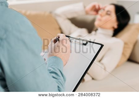 Cropped Shot Of Psychotherapist Writing On Clipboard And Young Woman Lying On Couch In Office
