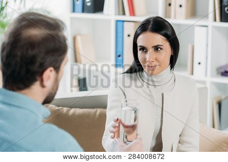 Young Woman Holding Glass Of Water And Looking At Psychotherapist In Office