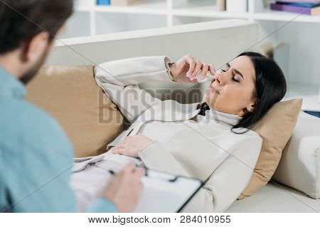 Selective Focus Of Psychotherapist Writing On Clipboard And Young Patient Crying On Couch