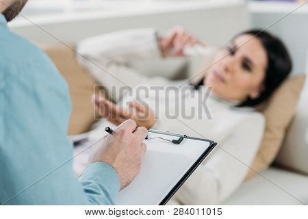 Cropped Shot Of Psychotherapist Writing On Clipboard And Young Patient In Depression Lying On Couch