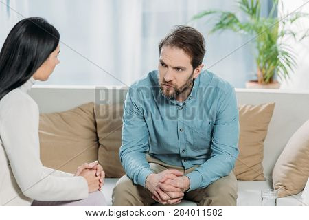 Young Female Psychotherapist And Upset Bearded Man Sitting And Looking At Each Other