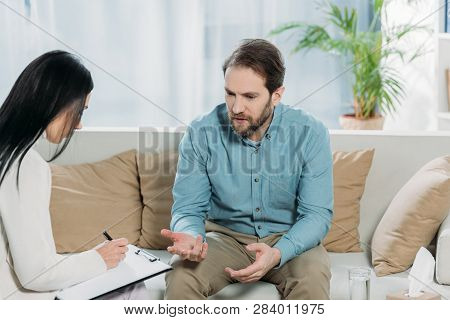 Psychotherapist Writing On Clipboard And Unhappy Bearded Man Sitting On Couch And Talking