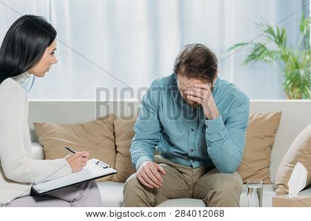 Young Psychotherapist Writing On Clipboard And Looking At Upset Bearded Man Sitting On Couch