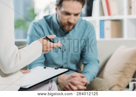 Cropped Shot Of Psychotherapist Holding Pen And Clipboard While Upset Bearded Man Sitting On Couch