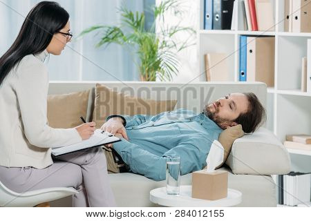 Young Psychotherapist In Eyeglasses Writing On Clipboard And Looking At Upset Bearded Man Lying On C