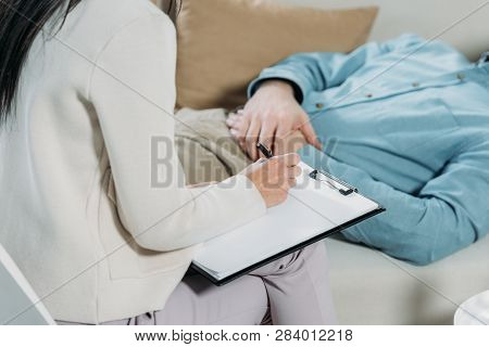 Cropped Shot Of Psychotherapist Writing On Clipboard And Male Patient Lying On Couch