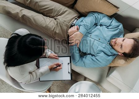 Overhead View Of Psychotherapist Writing On Clipboard And Male Patient Lying On Couch