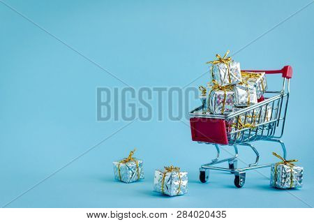 Small Shopping Cart With Presents On Blue Background. Trolley From A Supermarket Full Of Silver Gift