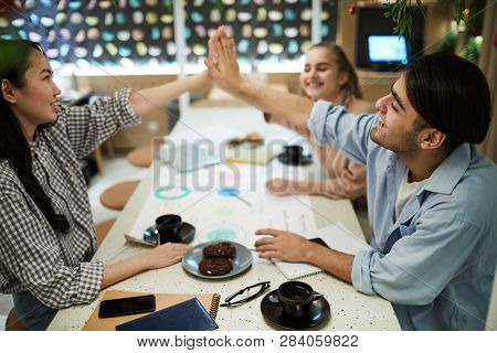 Cheerful Teenage Students Giving High Five To One Another Over Table With Cups Of Tea And Other Stuf