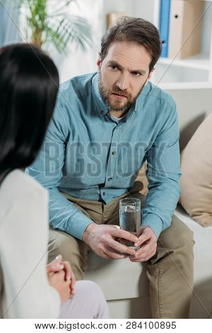 Cropped Shot Of Upset Bearded Man Holding Glass Of Water And Looking At Psychotherapist In Office