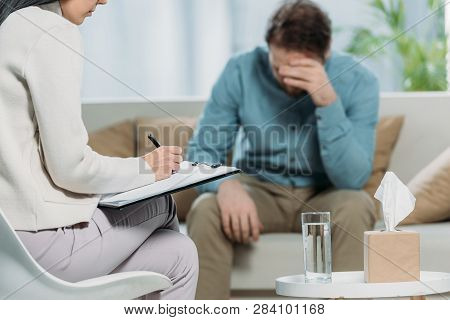 Cropped Shot Of Psychotherapist Writing On Clipboard And Upset Patient Sitting On Couch