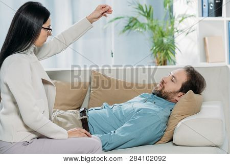 Side View Of Young Hypnotist With Pendulum And Bearded Man With Closed Eyes Lying In Couch During Hy