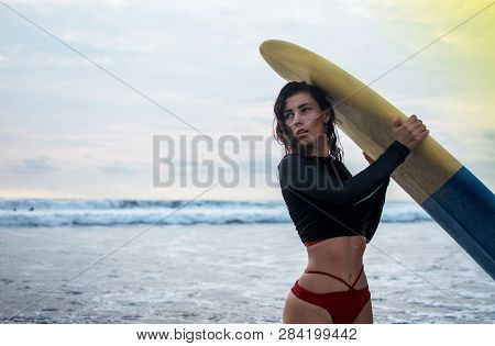 Outdoor Shot Of Young Girl Waxes Surfboard For Safe Riding Waves, Dressed In Black Swimsuit, Takes C