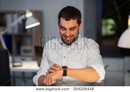 business, technology and time management concept - happy smiling businessman with smart watch at nigh office