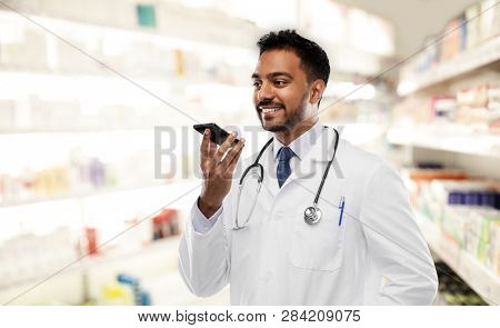 medicine, technology and healthcare concept - smiling indian male doctor or pharmacist in white coat with stethoscope using voice command recorder over drugstore background