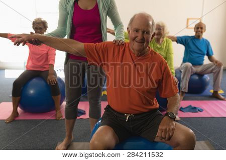 Front view of young female trainer assisting senior man making exercices on exercice ball in fitness studio