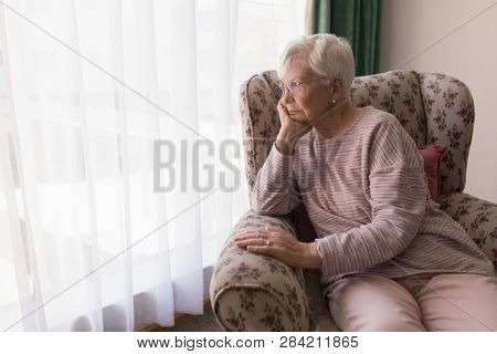 Front view of thoughting senior woman sitting on the couch and looking outside through window at home