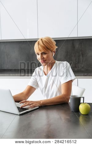 Smiling woman casualy dressed using laptop computer while sitting on a kitchen, wearing earphones