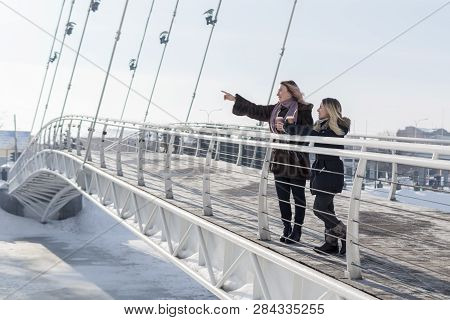 Woman Points Her Hand Into The Distance, Standing With A Friend On A Pedestrian Bridge In Winter