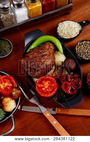 fresh roast beef meat rib eye steak with tomatoes,potatoes, hot chili pepper gern and pink peppercorn saucer, cutlery, set of spice, and red wine glass on light walnut table