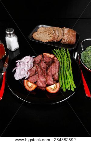business lunch fresh roast beef meat slices on black plate wotj cutlery asparagus boiled broccoli rye bun on wooden table