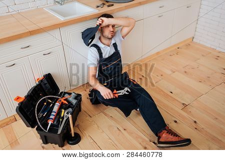 Tired Exhausted Plumber Sit On Floor In Kitchen. He Hold Hand On Forehead. Toolbox With Wrech And Ot