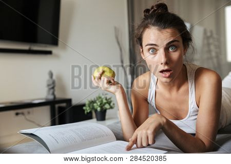 Beautiful young woman realxing on a couch at home, reading, eating green apple