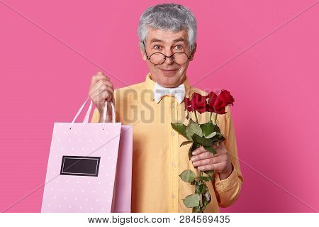 Photo Of Attractive Senior Man Looks Through Round Spectacles, Wears Formal Shirt And Bowtie, Holds 