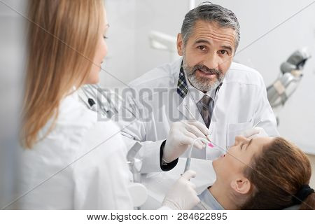 Woman With Opened Mouth Lying In Dentist Chair. Handsome Bearded Dentist Looking At Camera, Posing. 
