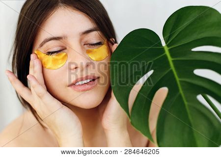 Portrait Of Beautiful Young Woman Applying Golden Eye Patches At Green Monstera Leaf On White. Happy