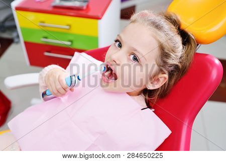 Baby Little Cute Girl Without Front Milk Teeth In Red Dental Chair With Electric Automatic Toothbrus